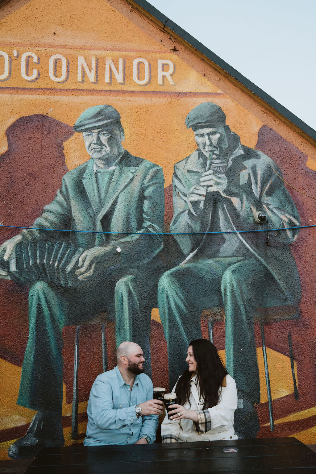 Jaime and Nolan clinking pints beneath the musicians mural